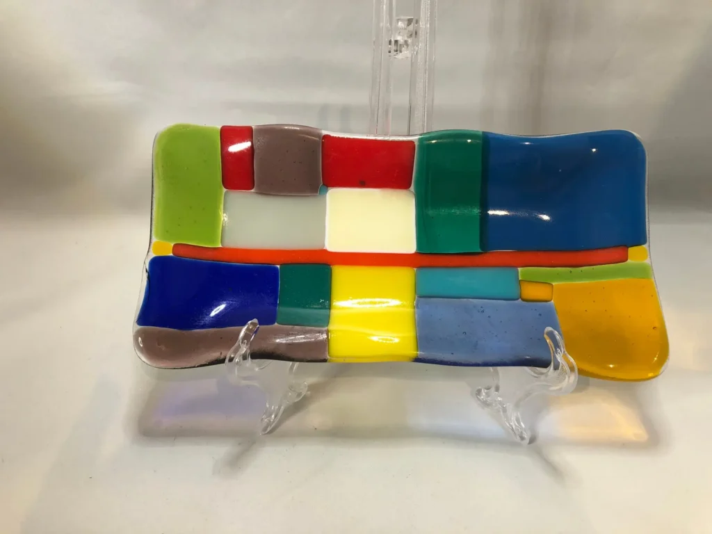 The 4 x 8 rectangular glass dish showcases a vibrant mosaic of red, blue, green, yellow, white, and gray squares and rectangles. It is displayed on a clear stand against a white background.