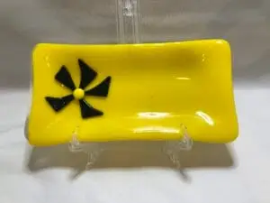 A 4 x 8 yellow glass dish features a decorative black pinwheel design on the left side. It is displayed on a clear stand against a plain background.