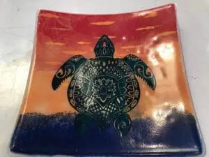 The Green Turtle 6 x 6 Dish showcases a detailed green sea turtle design set against an orange and red gradient sunset sky with small clouds, transitioning into a dark blue sea at the bottom.