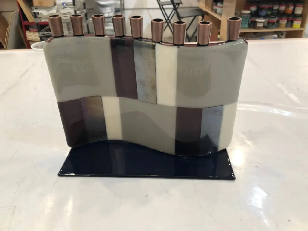 A modern menorah from the Menorahs collection features a wavy ceramic base with gray, cream, and brown rectangles on a dark glossy platform. Copper candle holders line the top, with shelves of paint cans visible behind it.