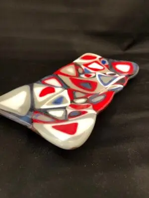 The 4 x 8 Dish is a colorful glass art piece with abstract red, white, and blue shapes on a dark background. The overlapping irregular shapes create a dynamic and vibrant design.