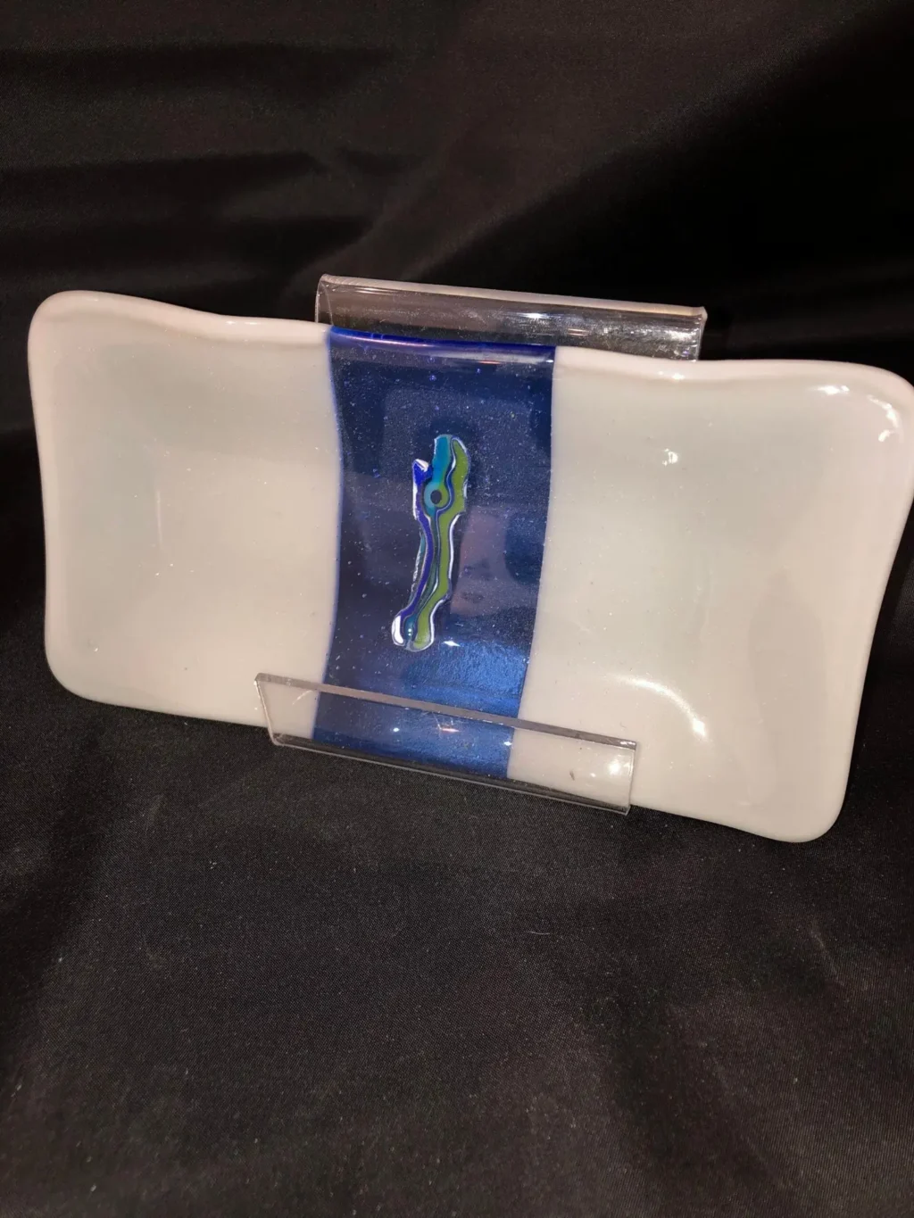 A 4 x 8 glass dish on a stand includes a smooth white surface with a bold, vertical blue stripe accented by a small, colorful abstract design, set against a dark fabric background.