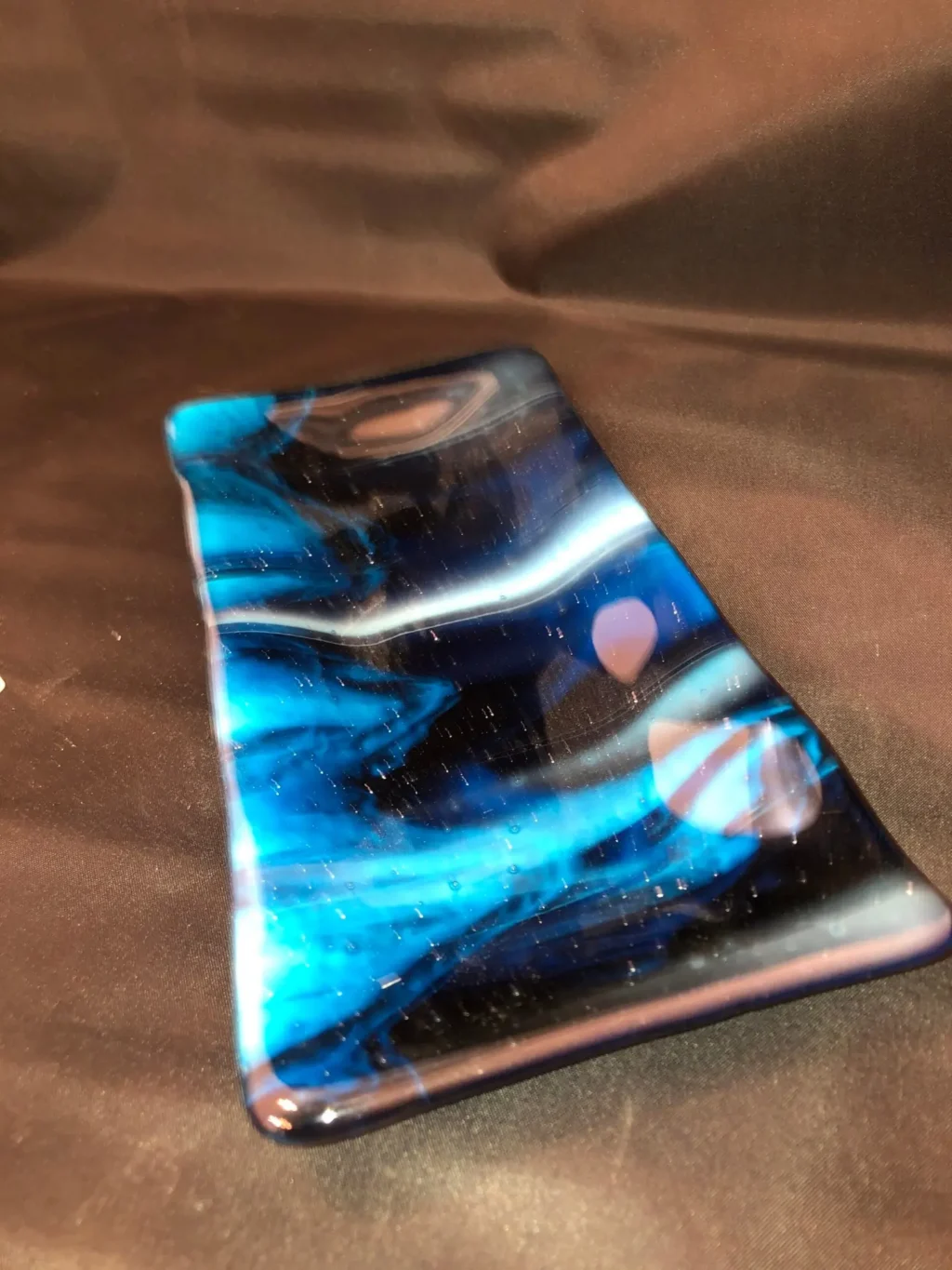 A 4 x 8 Dish with swirling blue, black, and white patterns rests on a dark fabric background, with light reflections creating shimmering effects.