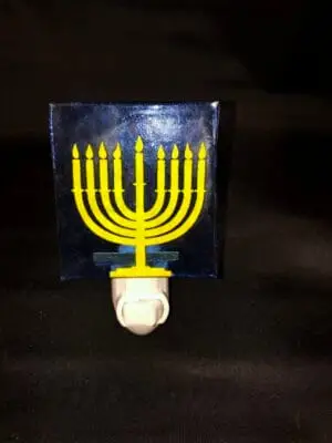 A nightlight with a yellow menorah showcasing nine lit candles on a dark backdrop, mounted on a square transparent panel and attached to a white base. Product: Nightlights.