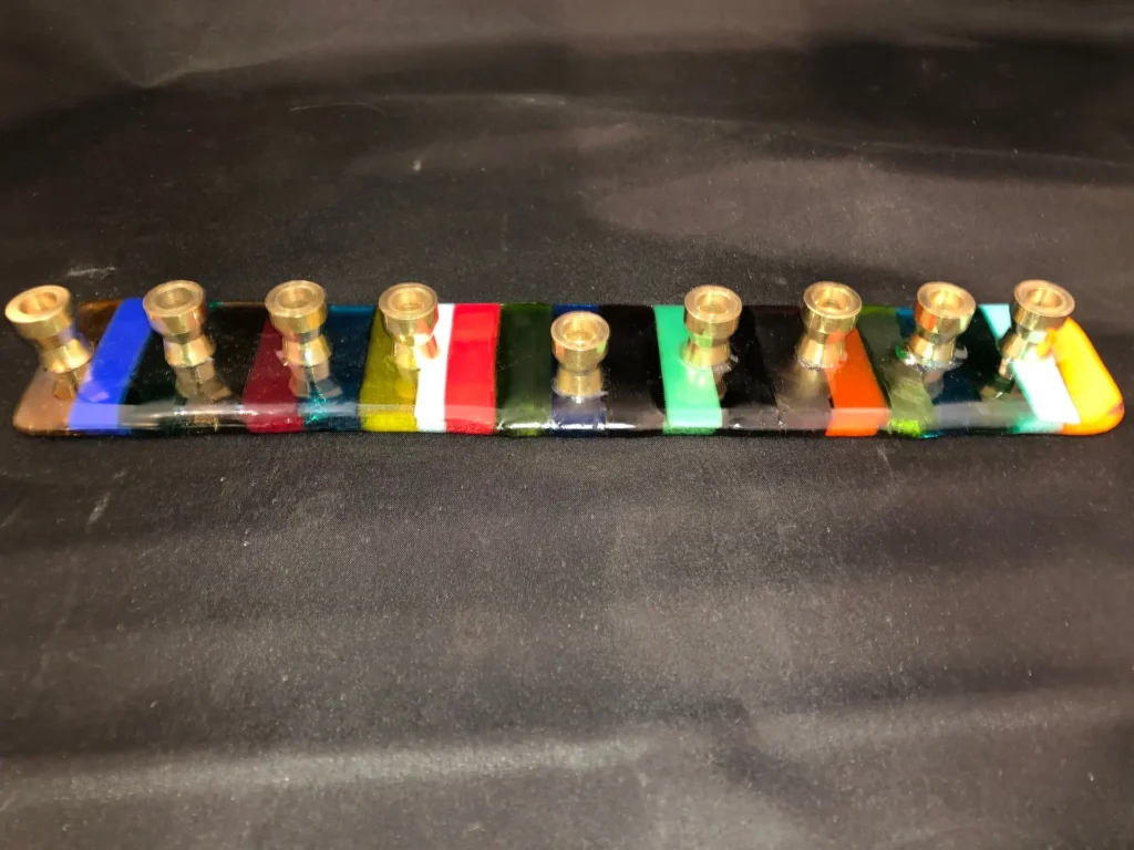 A glossy-finished multicolored Menorah features nine gold candle holders on a rectangular base, showcasing colorful segments in blue, yellow, red, green, and black against a dark background.