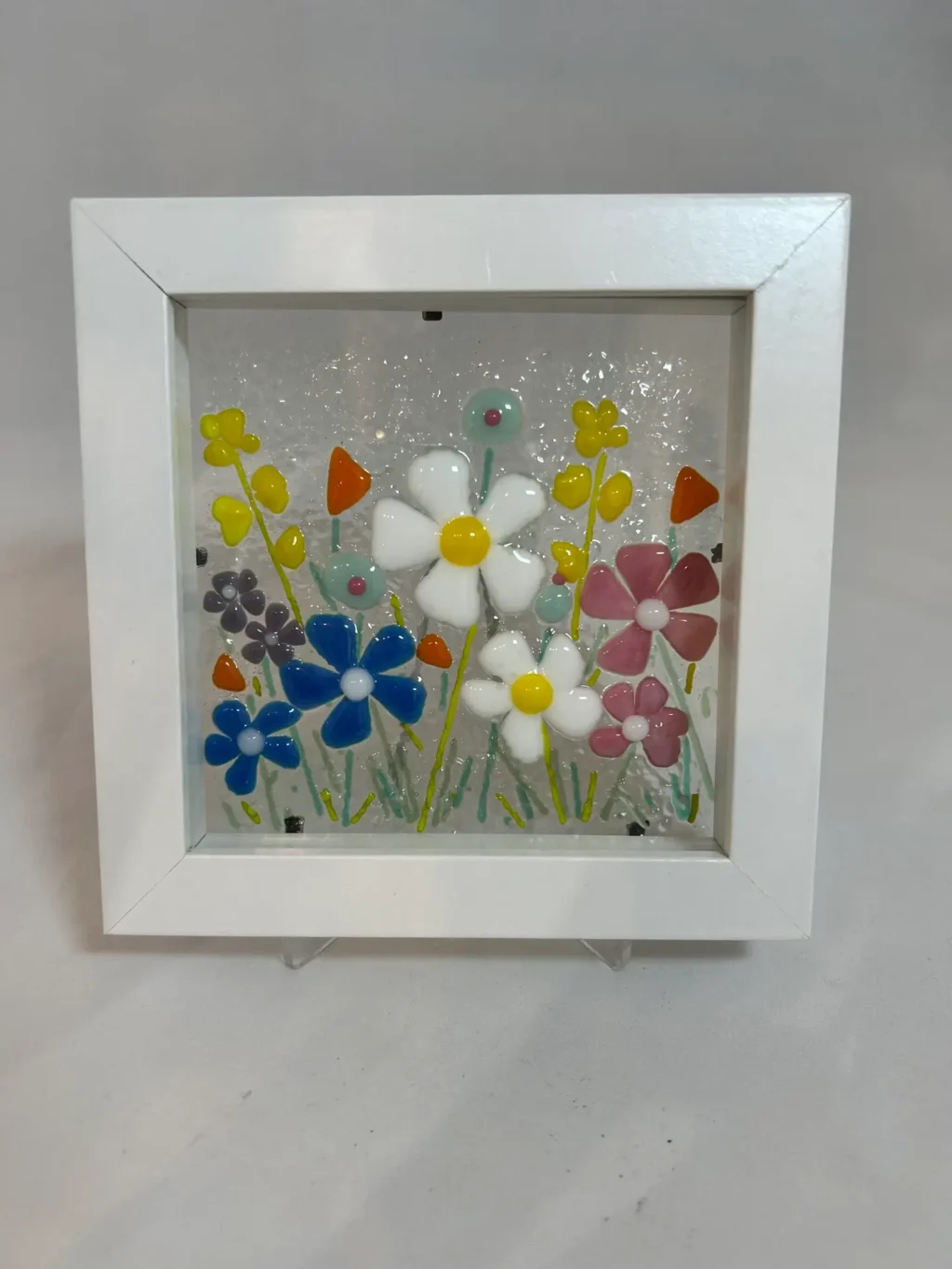 The Enchanted Garden is a framed decorative glass art piece showcasing vibrant white, blue, pink, and orange flowers.