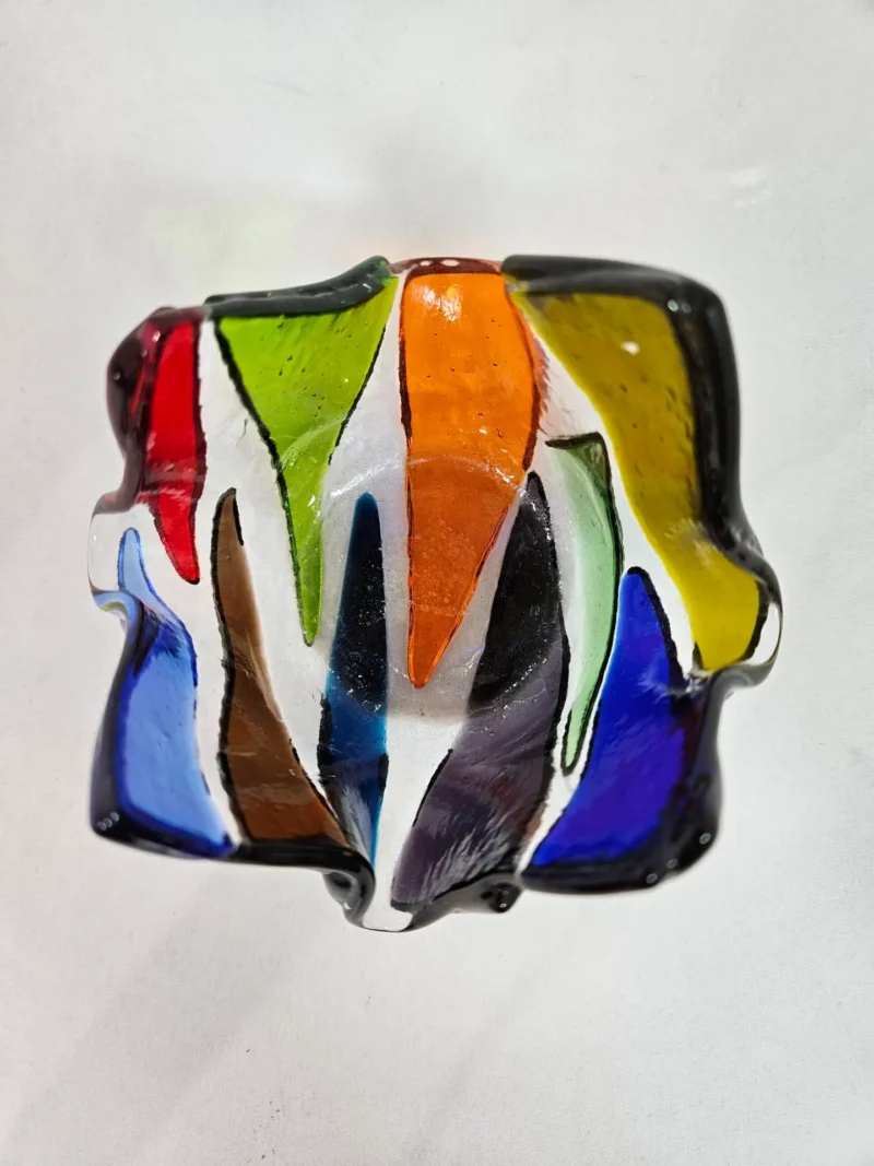 The Prism Flame is a colorful glass abstract sculpture featuring jagged shapes and a mix of red, green, orange, white, blue, and brown hues.