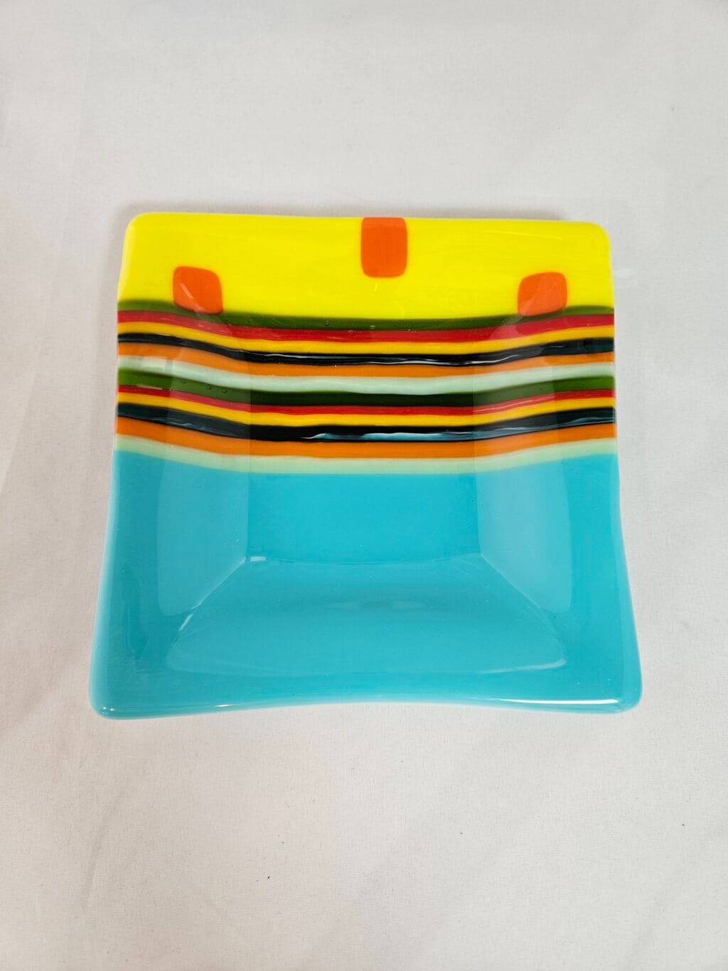 The Striped Radiance plate is a decorative square with a colorful design: the top section features yellow with three orange squares, followed by black, green, yellow, and red stripes. The bottom half is solid light blue. It rests on a plain white surface.