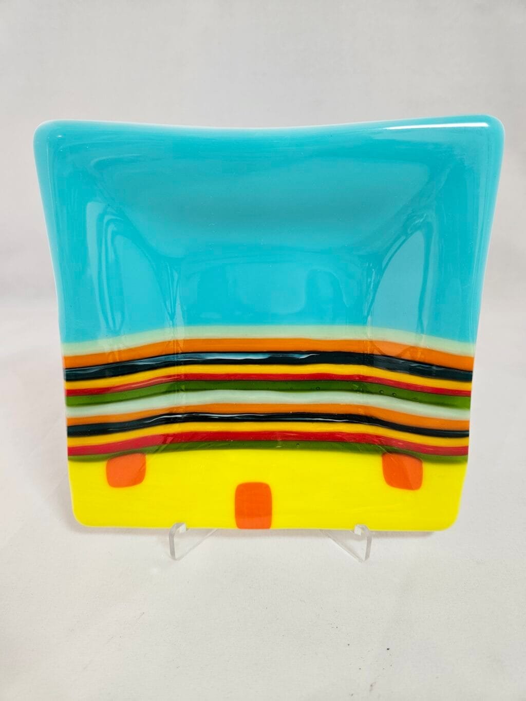 The Striped Radiance plate is a square, fused glass design with a turquoise-to-yellow gradient and horizontal stripes in black, red, brown, white, and green. Two red squares adorn the lower yellow section. It boasts a glossy finish and is currently on display.