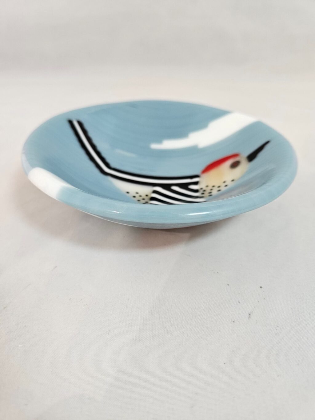 The Skybound Woodpecker is a round fused glass dish featuring an artistic bird design with a red head, white body, and black spots, against a light blue backdrop with cloud-like shapes. The birds wings are outstretched.