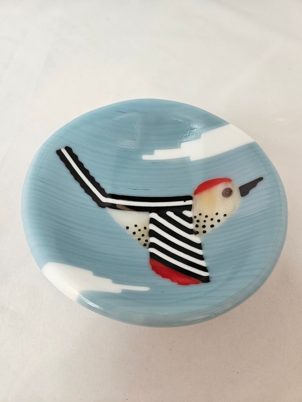 The Skybound Woodpecker is a round fused glass dish featuring an artistic bird design with a red head, white body, and black spots, against a light blue backdrop with cloud-like shapes. The birds wings are outstretched.