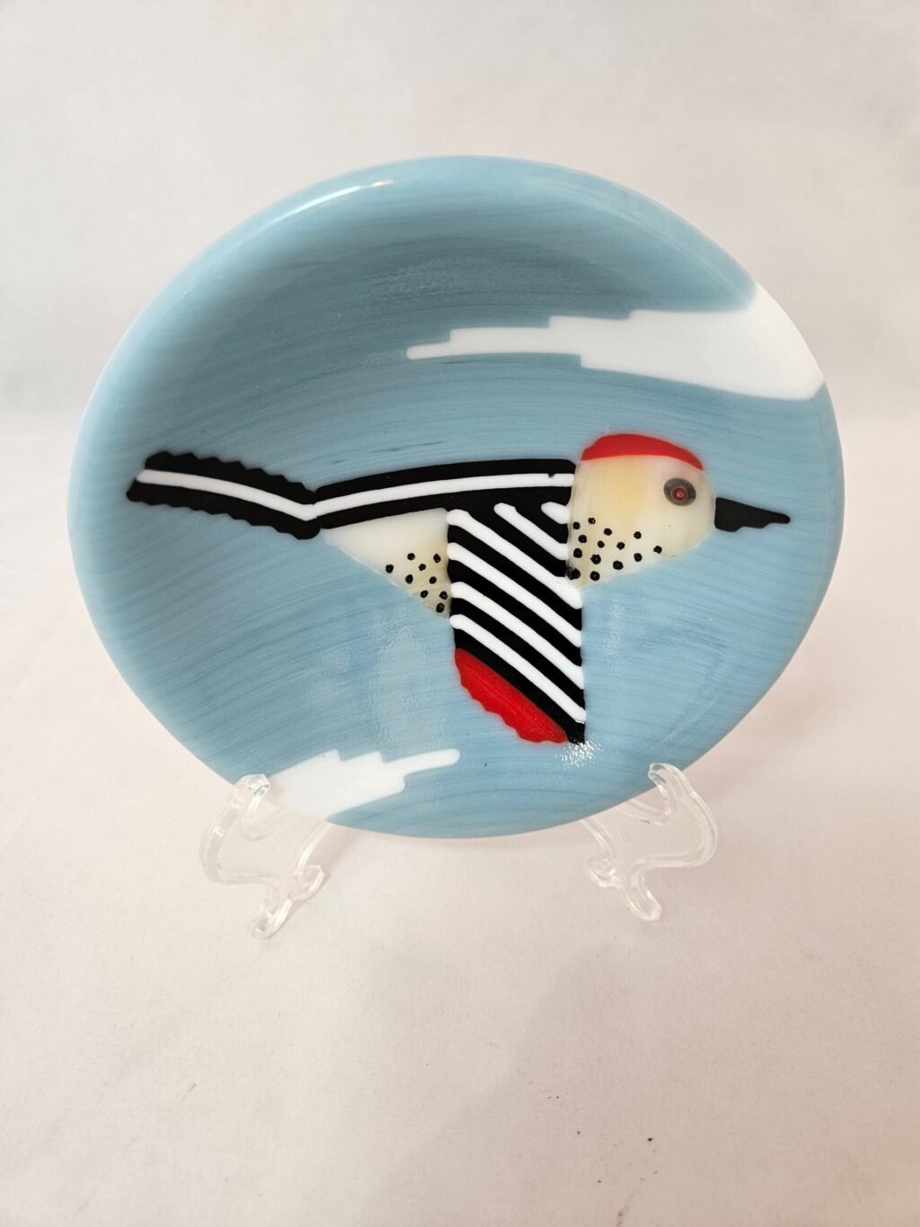 The Skybound Woodpecker is a round fused glass dish featuring an artistic bird design with a red head, white body, and black spots, against a light blue backdrop with cloud-like shapes. The birds wings are outstretched.