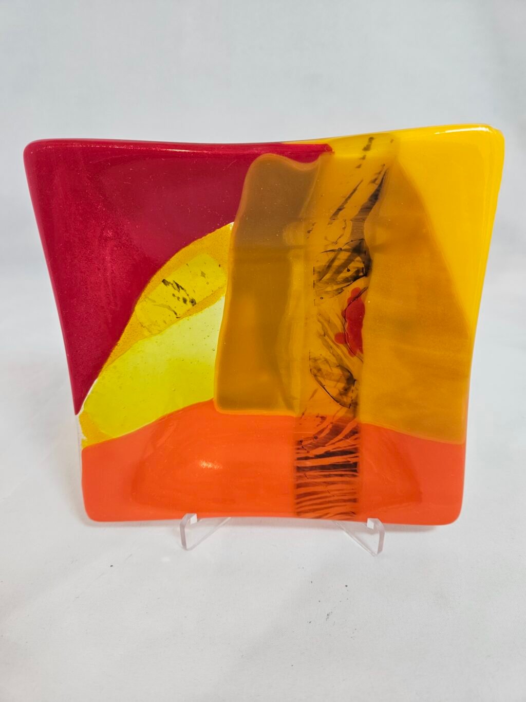 The Radiant Horizon is a square, abstract fused glass plate with vibrant red, orange, yellow and brown. Its colors blend dynamically on its slightly curved surface.