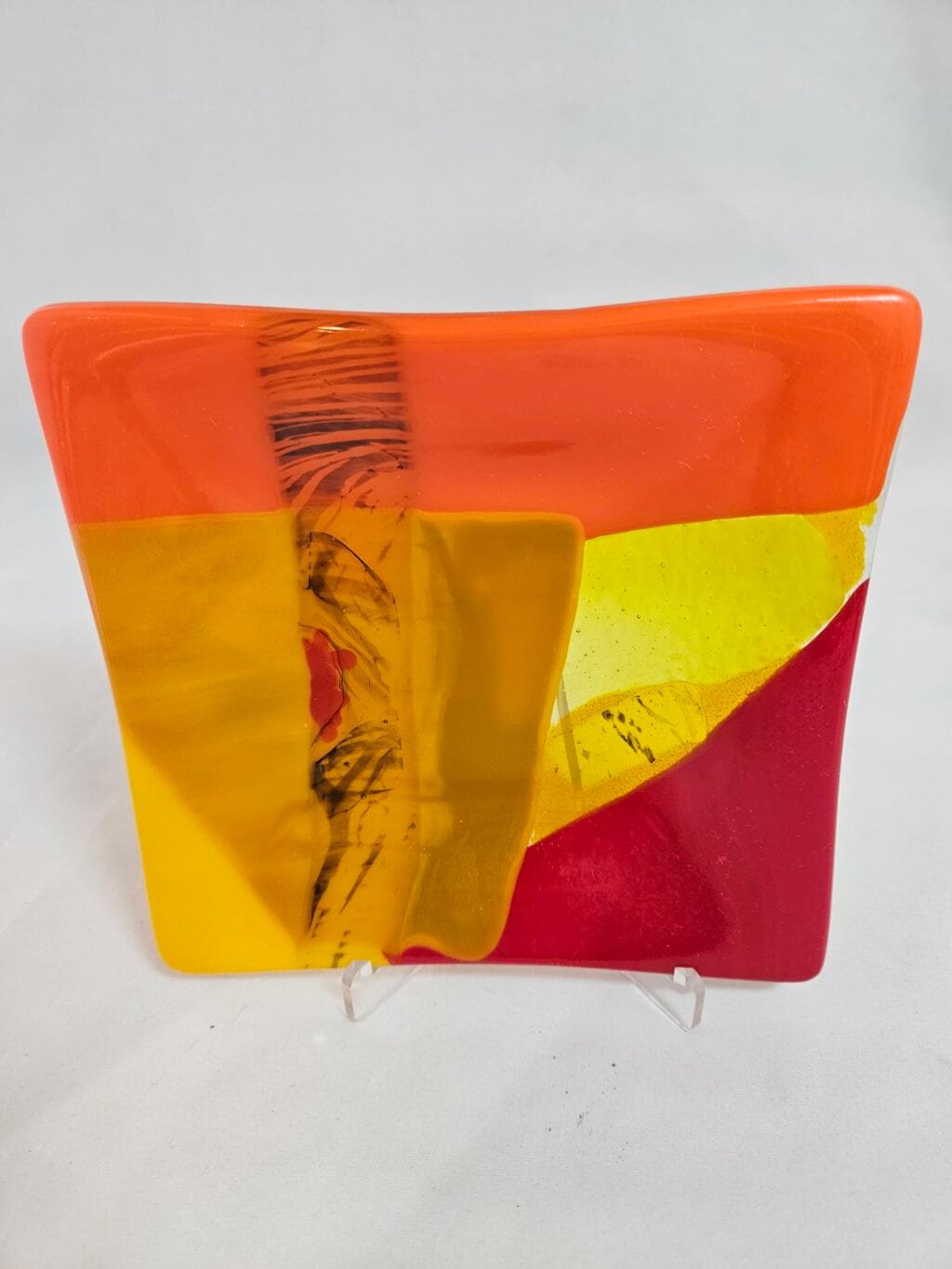 The Radiant Horizon is a square glass plate with vibrant abstract patterns of orange, yellow, and red. A central vertical streak shows black and orange markings against a white background. It is elegantly displayed on a clear stand.