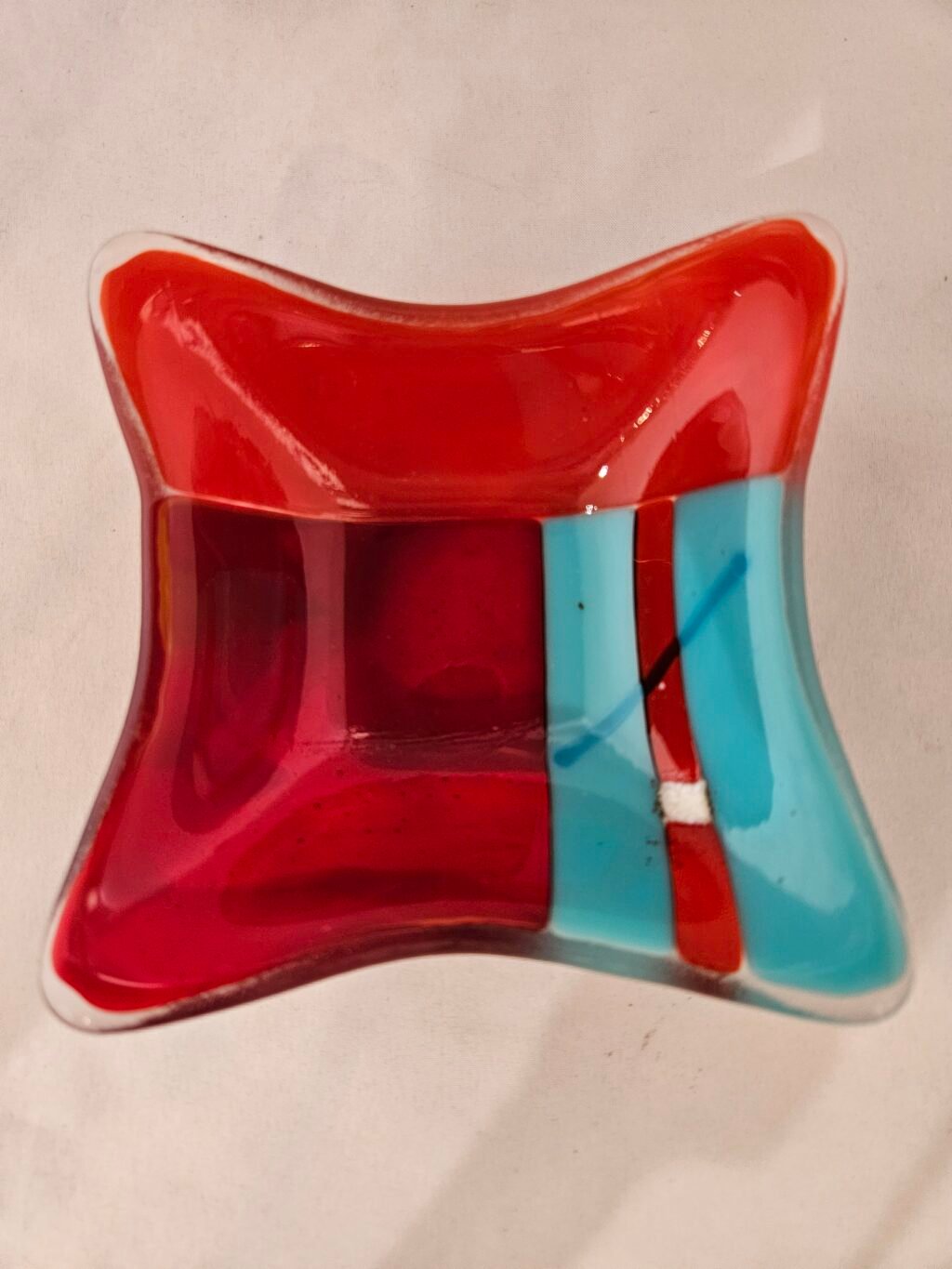 The Fiery Contrast dish is a square fused glass piece with wavy edges, showcasing bold red, turquoise, and orange sections divided by thin black lines.