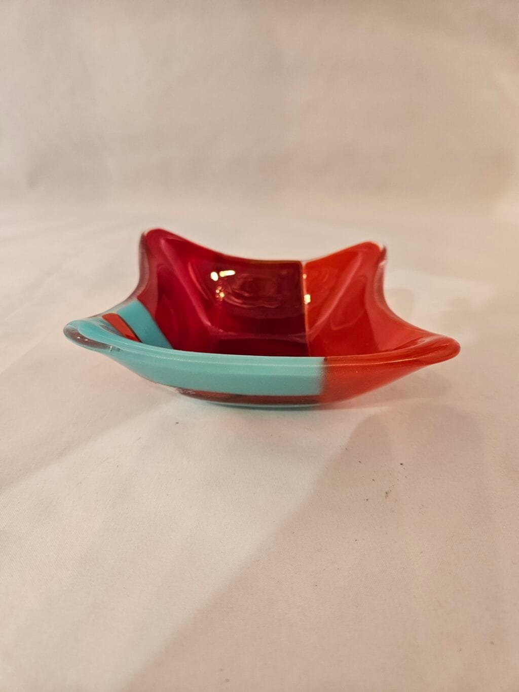 The Fiery Contrast dish is a square fused glass piece with wavy edges, showcasing bold red, turquoise, and orange sections divided by thin black lines.