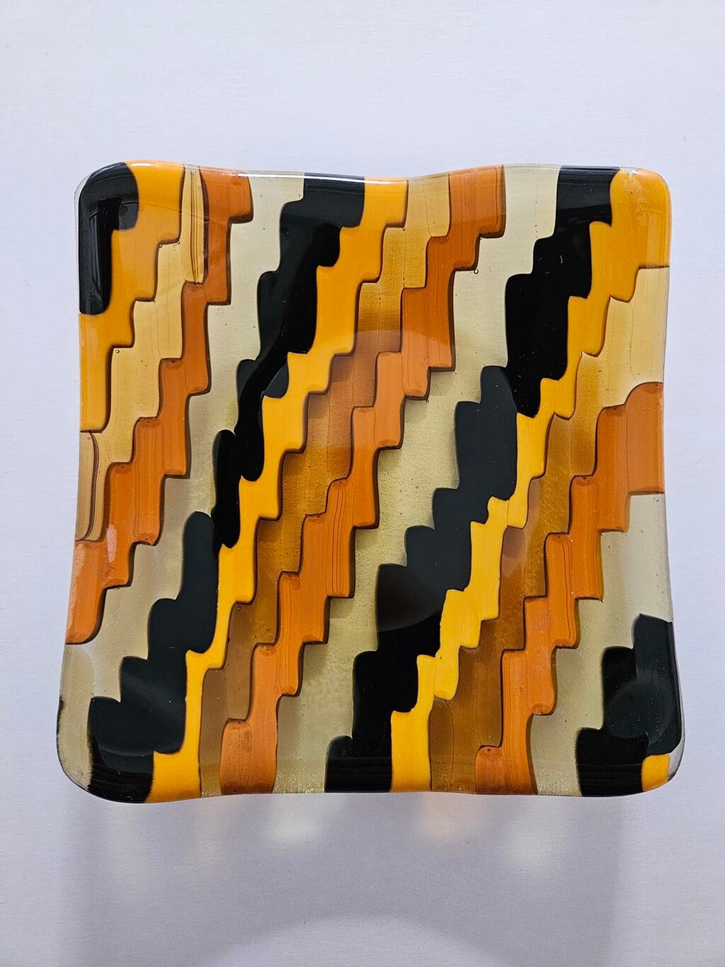 Introducing Emberwave: a square fused glass dish featuring wavy patterns in alternating shades of orange, amber and black. It creates a striking zigzag effect on the surface and includes a slightly wavy edge.