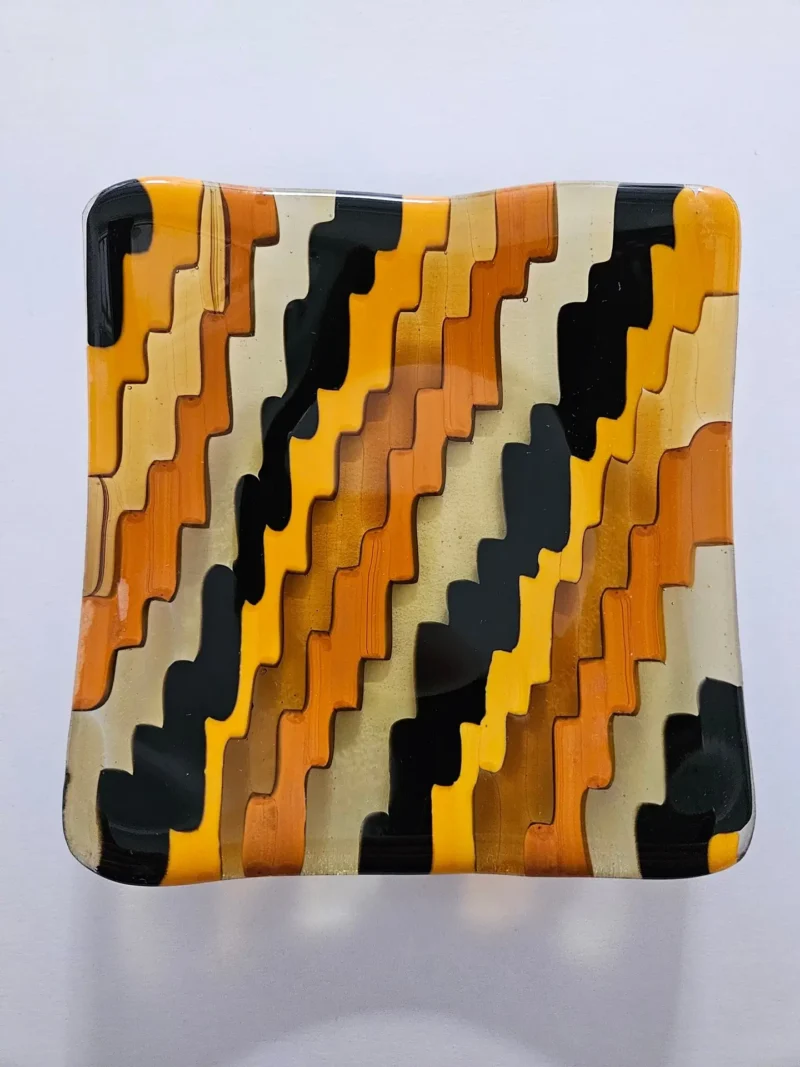 Introducing Emberwave: a square fused glass dish featuring wavy patterns in alternating shades of orange, amber and black. It creates a striking zigzag effect on the surface and includes a slightly wavy edge.