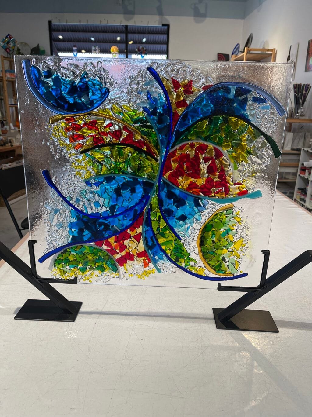 Certainly! Here is your description with the background, surface, stand, position, and things near or beside the product removed: The Joy In Motion fused glass panel features curved blue lines and vibrant red, yellow, green, and blue fragments. It exemplifies contemporary glass art for any studio setting.