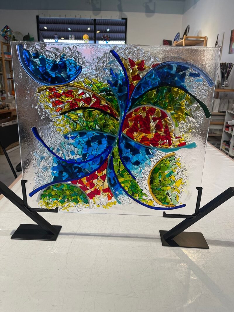 Certainly! Here is your description with the background, surface, stand, position, and things near or beside the product removed: The Joy In Motion fused glass panel features curved blue lines and vibrant red, yellow, green, and blue fragments. It exemplifies contemporary glass art for any studio setting.