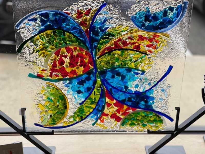 Certainly! Here is your text with the background, surface, stand, position, and things near or beside the product removed: The Joy In Motion fused glass sculpture features abstract colorful patterns in blue, red, yellow, and green on a square panel.
